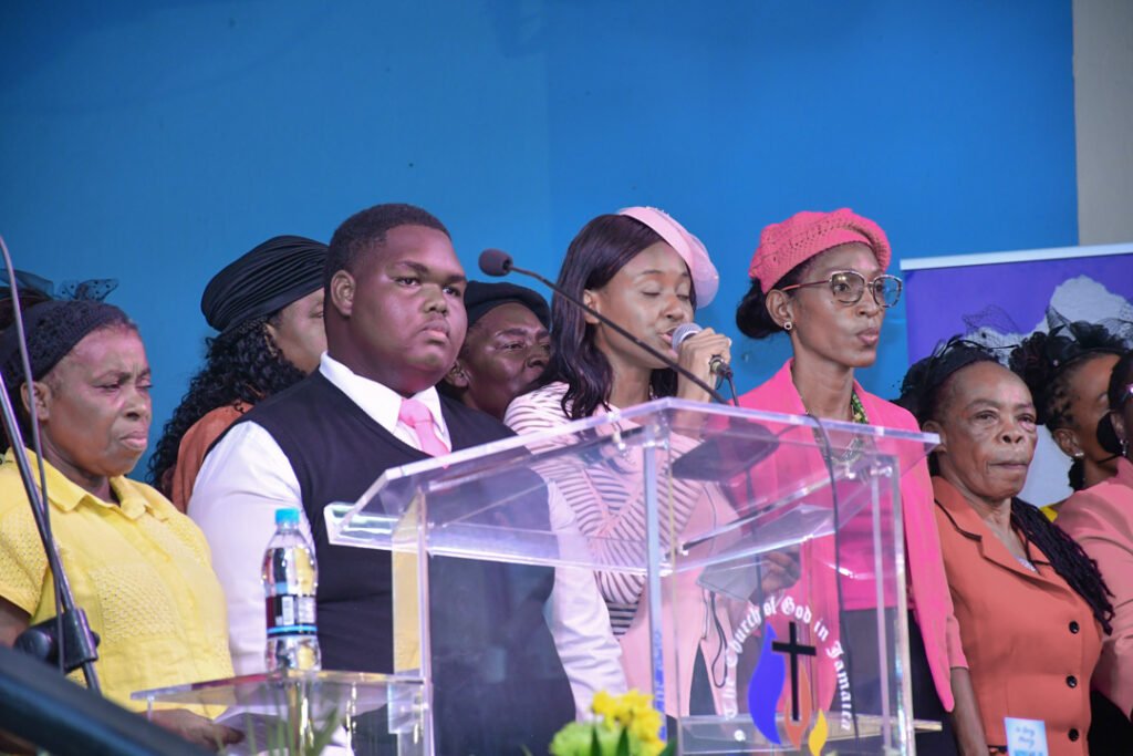 A group of people, including men and women, stand at a podium in a church setting. The individuals are dressed in various formal and colorful outfits, with some wearing hats and accessories. One person is speaking into a microphone while others appear to be listening attentively.