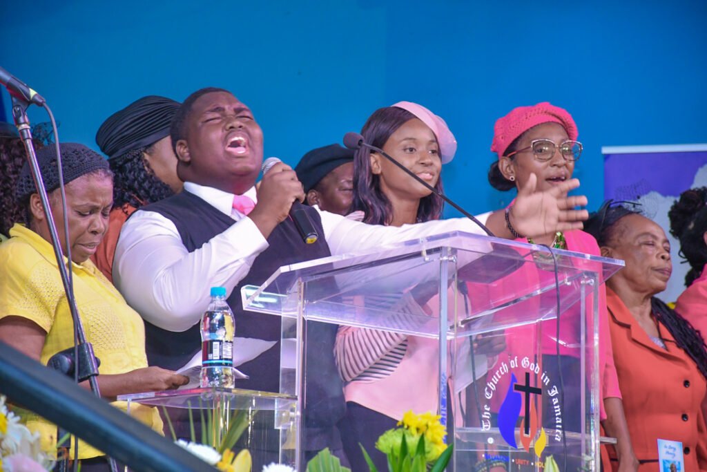 A group of people singing passionately at a church service, with a young man leading with a microphone and various attendees visibly engaged in worship.
