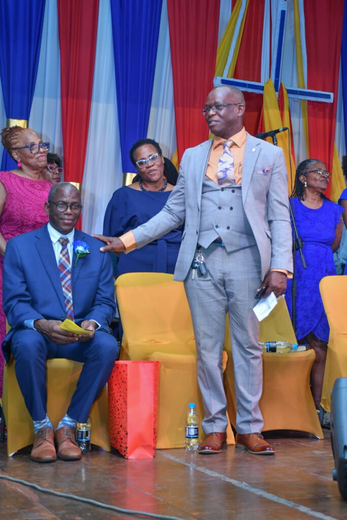 A group of people at a formal event, with a speaker engaging an audience member seated on stage. Colorful backdrops and decorations visible.