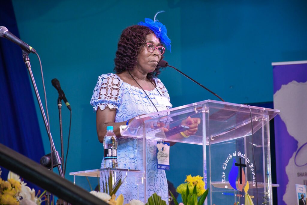 A woman speaking at a podium with a microphone, wearing a light blue dress and a blue hat, in a church setting with flowers on the table.