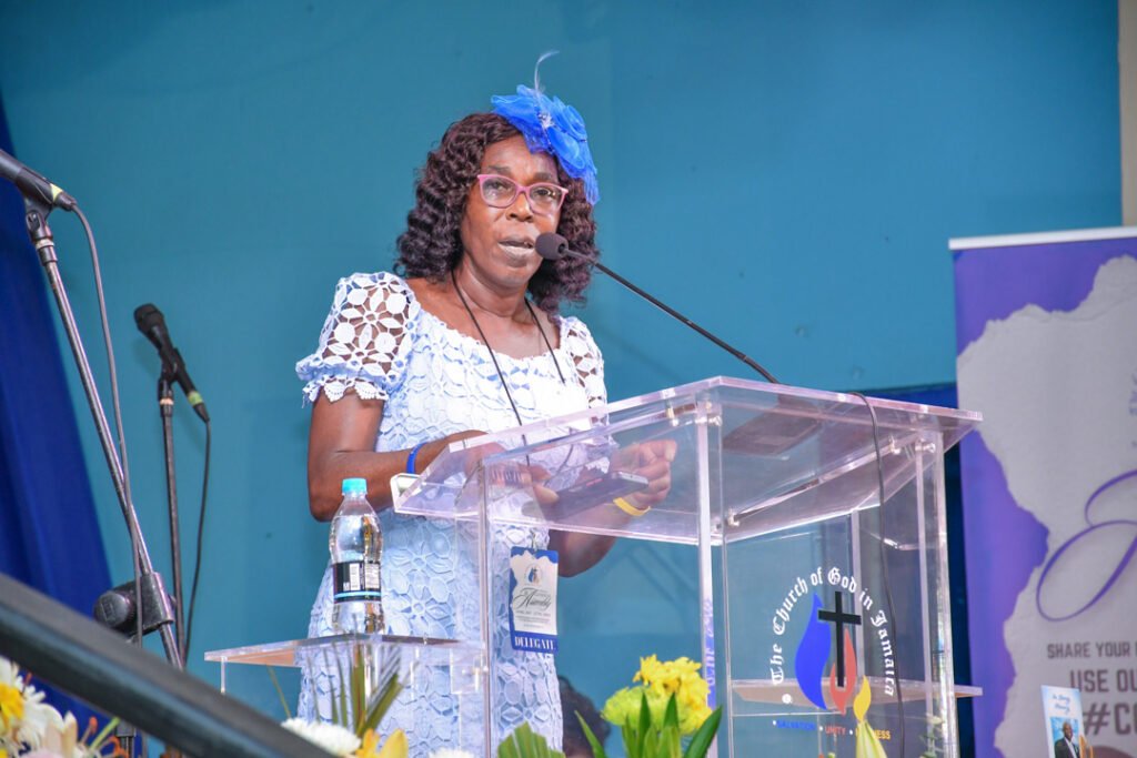 A woman speaking at a podium during an event, wearing a blue dress and a blue fascinator, with flowers and a water bottle on the podium.