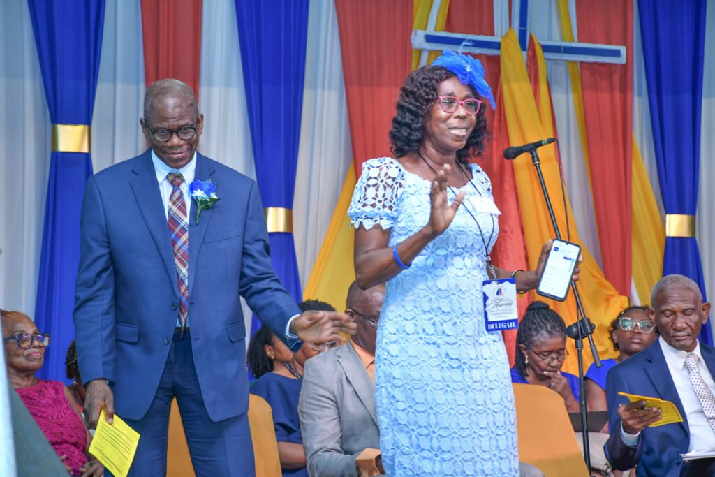 A well-dressed man and woman are engaged in conversation at an event. The man wears a blue suit with a patterned tie and glasses, while the woman is in a light blue dress with lace details, speaking into a microphone.
