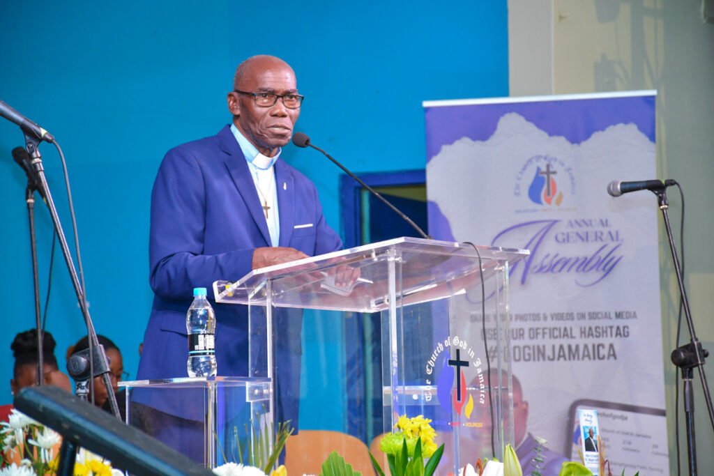 A speaker giving a presentation at a podium during an event, with a backdrop featuring the Church logo and details about the Annual General Assembly.