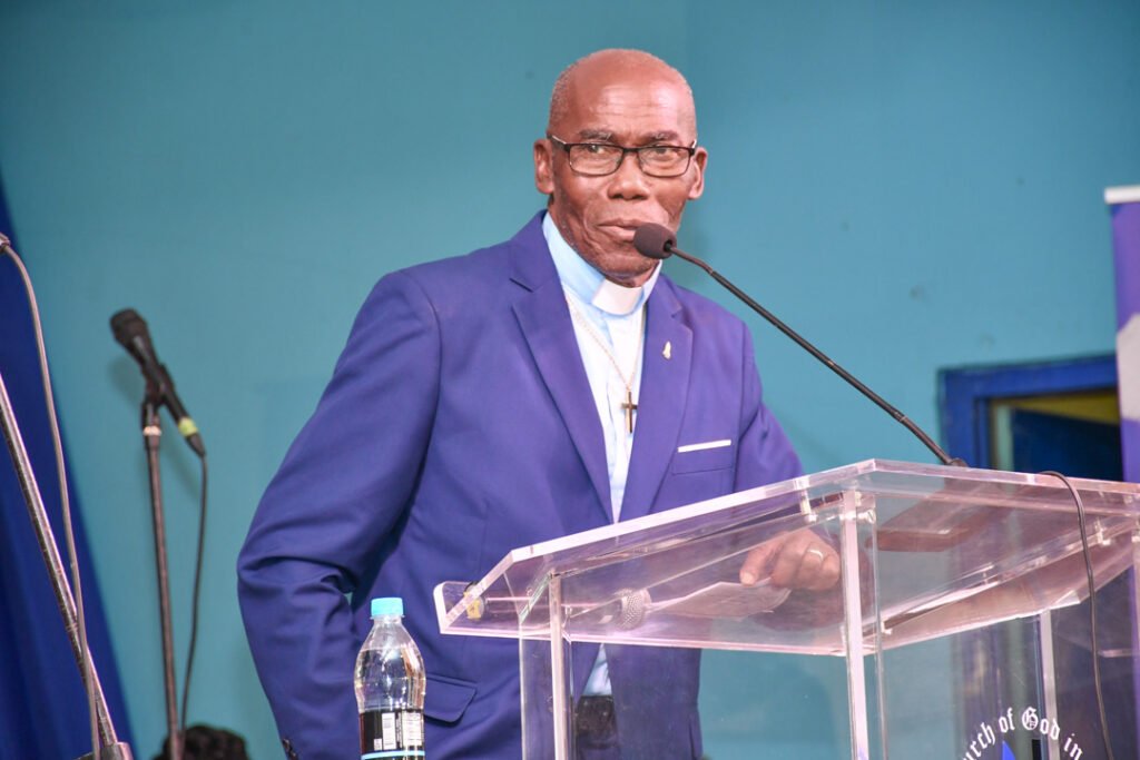 A speaker in a blue suit, wearing glasses and a clerical collar, stands at a podium with a clear backdrop. The setting appears to be a church or conference stage.