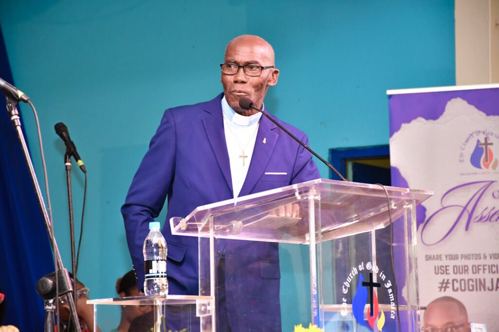 A speaker in a blue suit stands at a podium with a transparent lectern, addressing an audience. A microphone is set up nearby, and a banner with religious symbols is visible in the background.