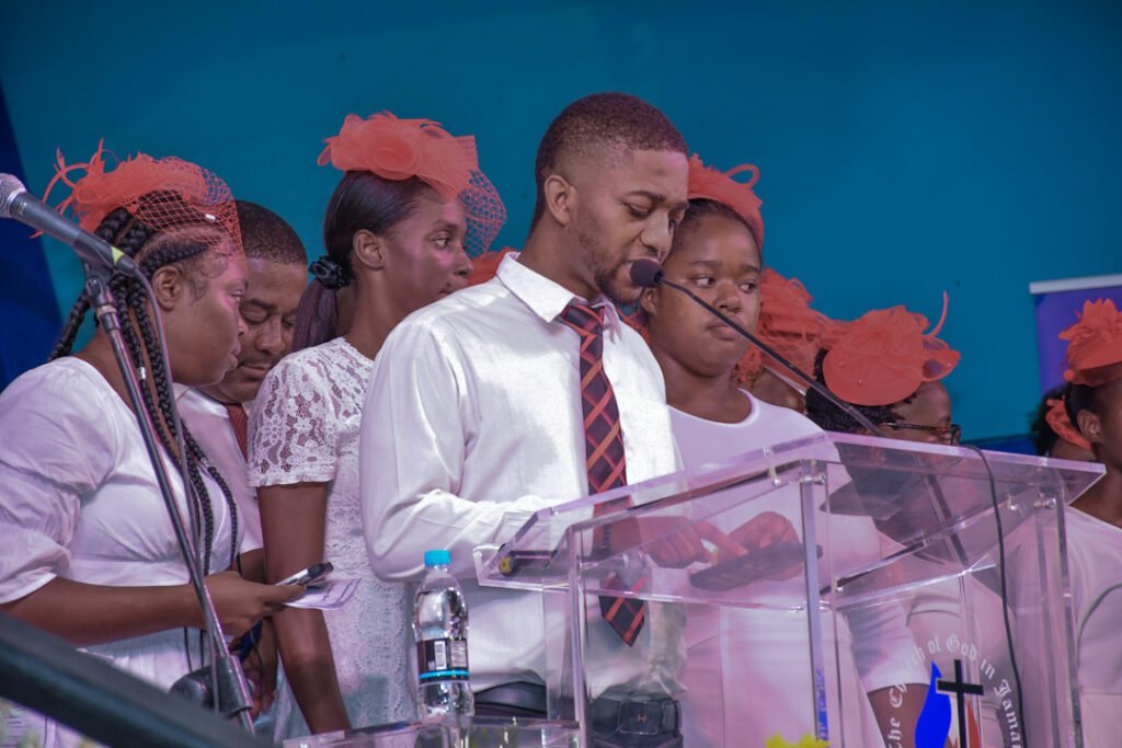 A group of individuals in formal attire, including white dresses and red hats, participates in a ceremony, with one person speaking at a podium.