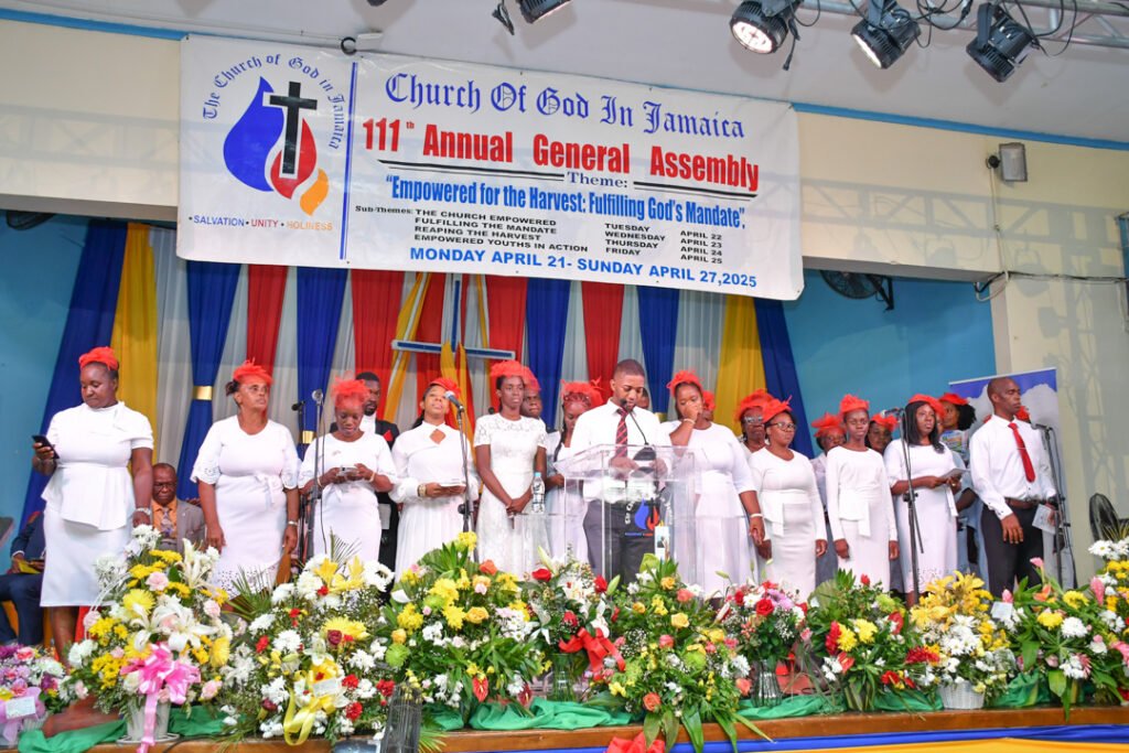Group of people on stage during a church event, wearing white attire and red accents, with a banner in the background displaying the theme of the assembly.