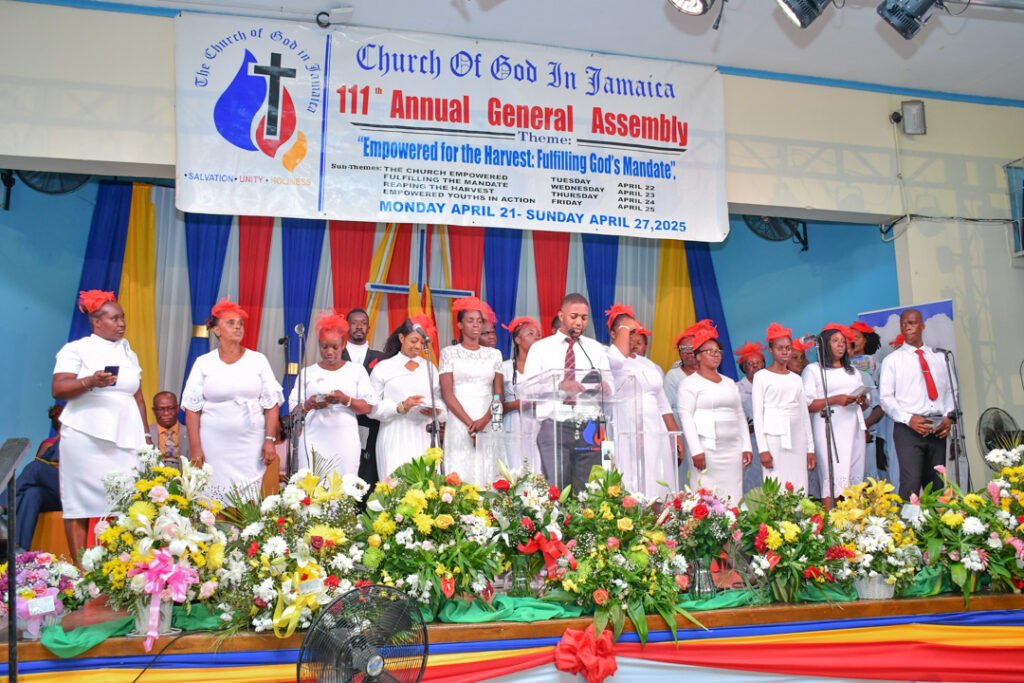 A group of people dressed in white attire with red accents stands on a stage, singing. They are surrounded by colorful decorations and flowers, with a banner in the background announcing the 111th Annual General Assembly of the Church of God in Jamaica, scheduled for April 21 to April 27, 2025.