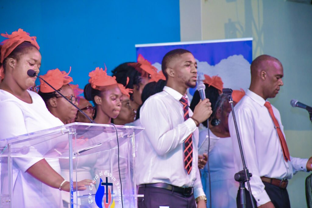A choir performing on stage, wearing white outfits and orange headpieces, with a male vocalist speaking into a microphone.