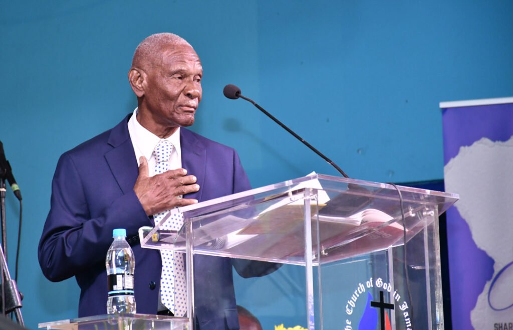 A man in a suit stands at a clear podium, speaking passionately. He has a serious expression and is gesturing with one hand. Behind him, there is a colorful backdrop with a map of Africa.