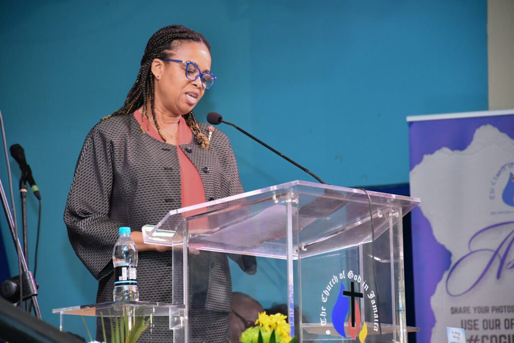 A woman with braided hair and glasses speaks at a podium during a church event, with floral arrangements and a banner in the background.