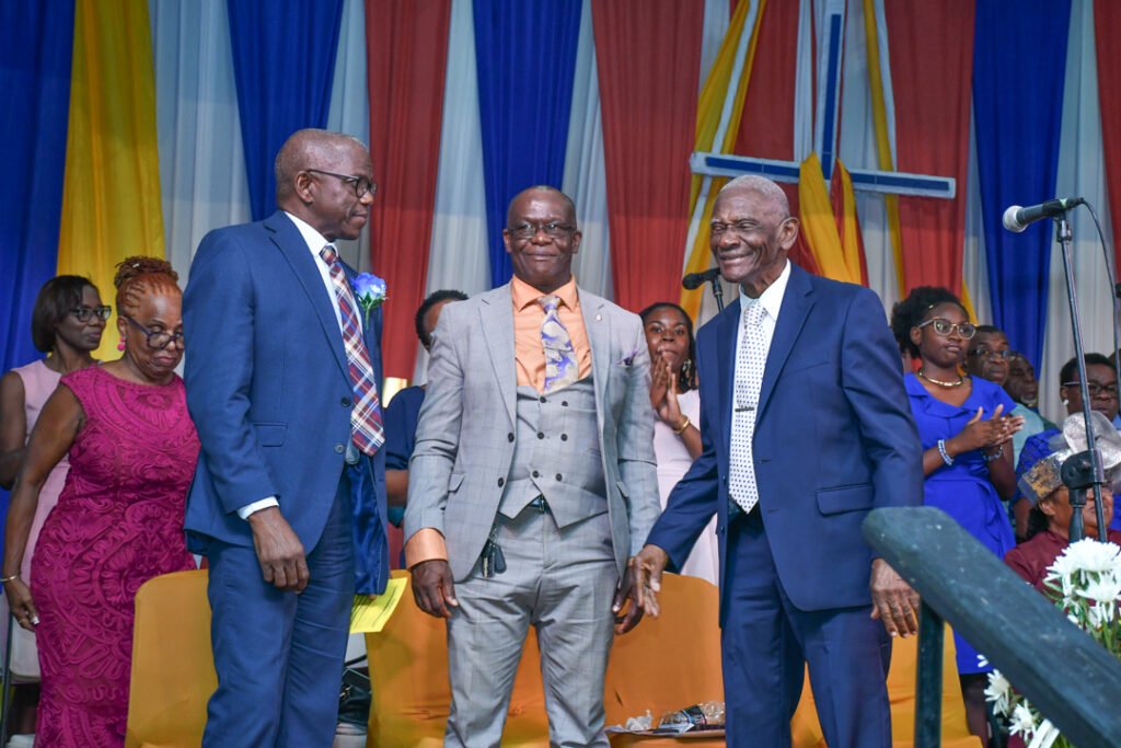 Three men in formal attire smiling and interacting during a celebratory event, with a colorful backdrop featuring drapes and a cross visible.