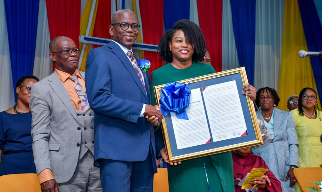 A formal event where a woman in a green dress receives an award from a man in a suit, both smiling. Other attendees are visible in the background.