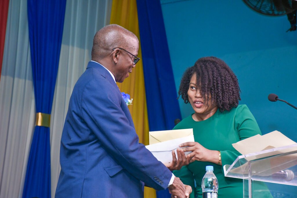 A man in a suit hands an envelope to a woman in a green dress during a formal event, with blue and yellow drapes in the background.