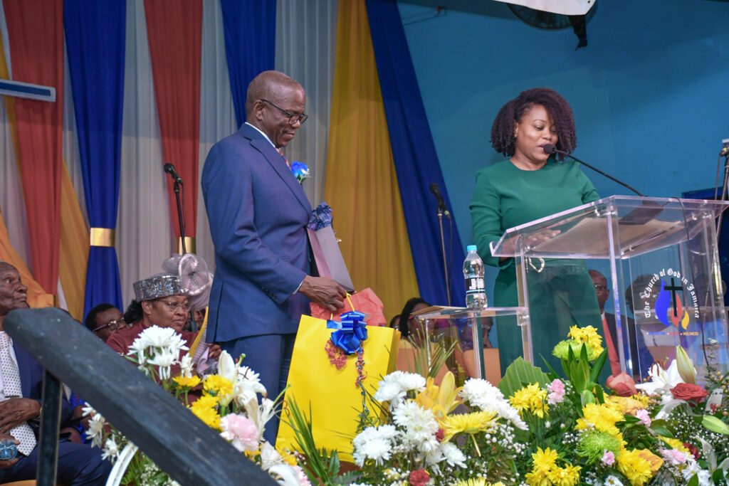 A man in a blue suit receives a gift bag from a woman in a green dress at a podium adorned with flowers during a celebration event.