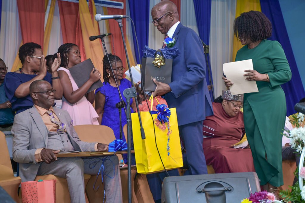 A group of people at an event, some clapping and holding documents, while a man in a blue suit receives a gift and a certificate from a woman in green. The backdrop features colorful drapes.