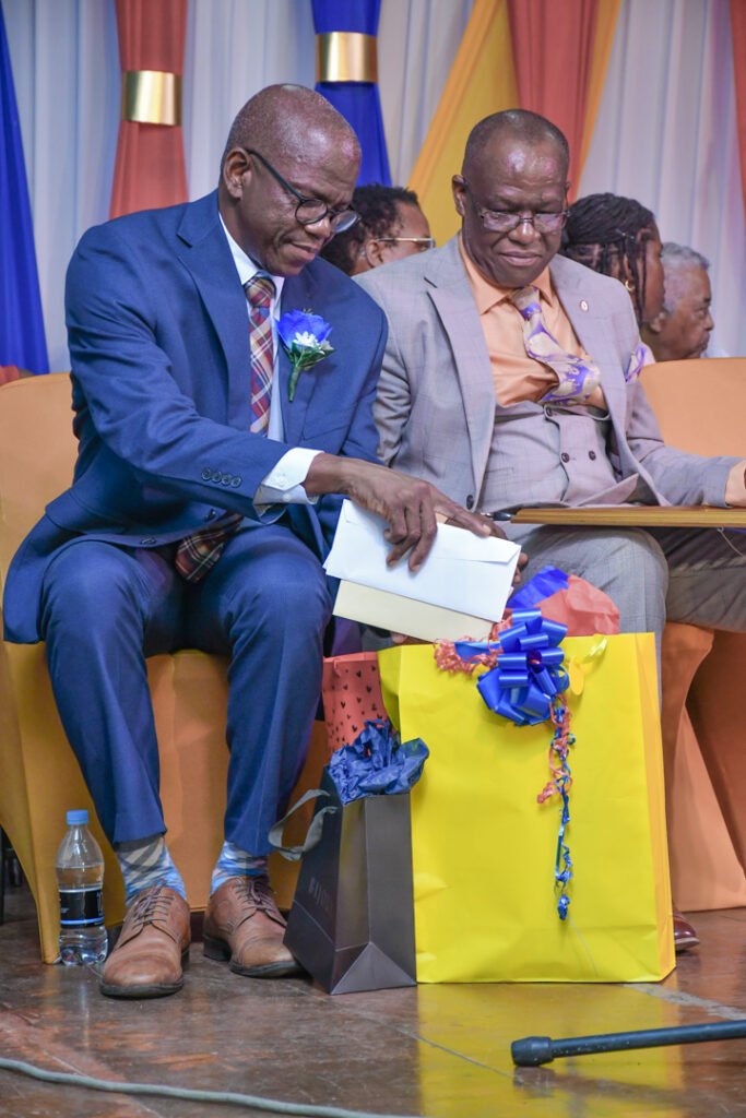 Two men seated on stage, one wearing a blue suit with a flower on the lapel, the other in a light gray suit, both holding gifts on their laps. A water bottle and a microphone are visible in the foreground.