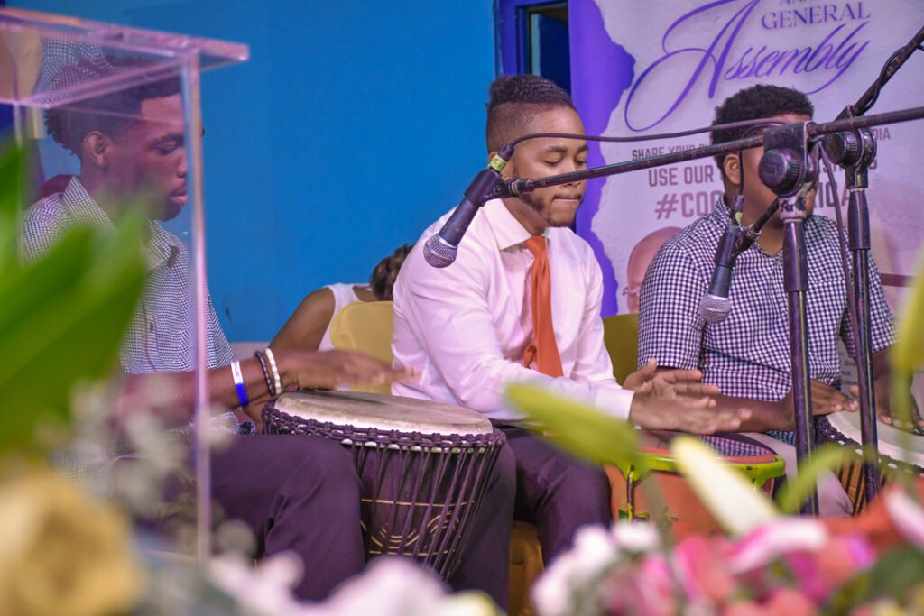 Three musicians playing percussion instruments at an event, with a microphone in front of them and floral decorations in the foreground.