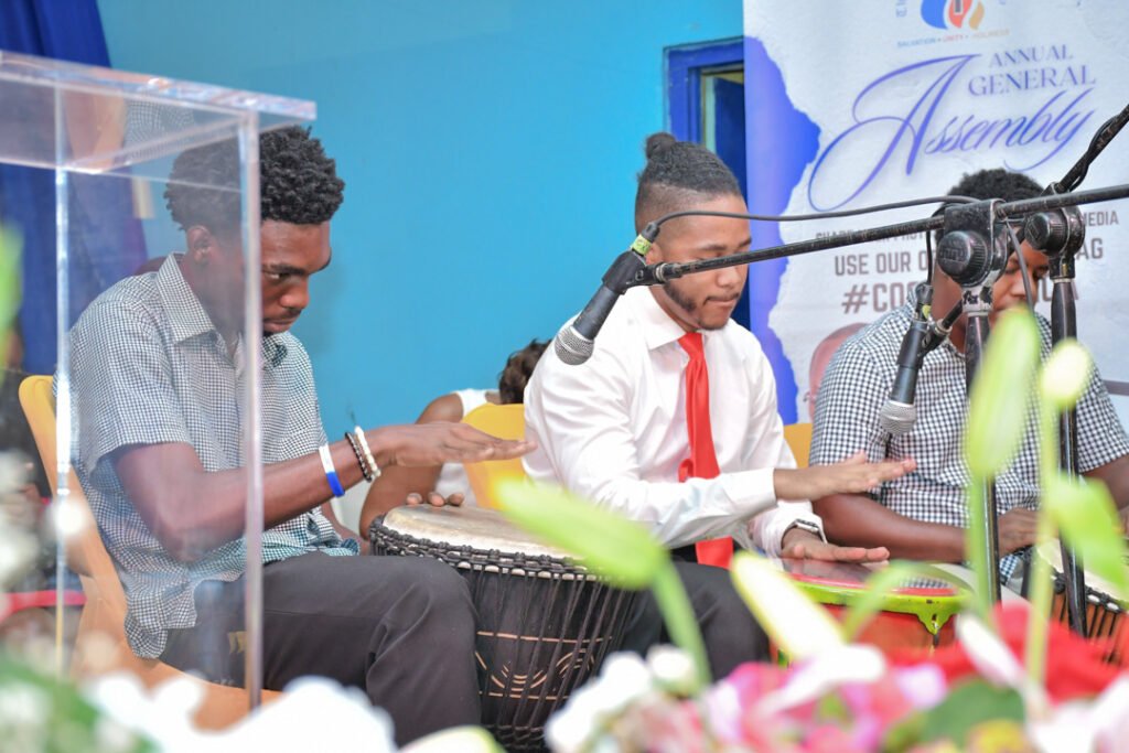 Three young men playing drums on stage during an event, with microphones positioned in front of them and a colorful backdrop.