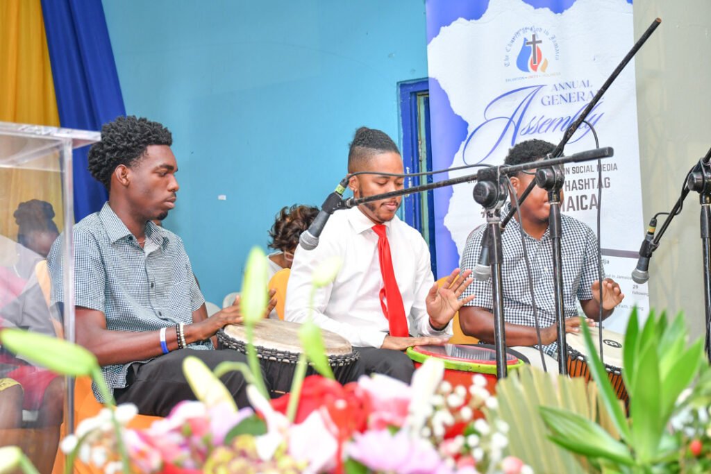 Three young men playing percussion instruments during an assembly, with a colorful backdrop and plants in the foreground.