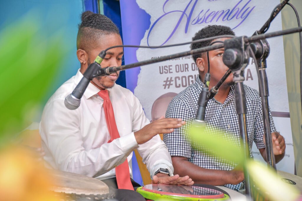 Two musicians performing at an event, one playing a drum and the other holding a microphone, with a colorful backdrop and greenery in the foreground.