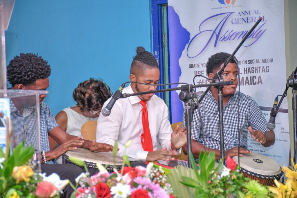 A group of young musicians playing percussion instruments at a cultural event, with a vibrant floral arrangement in the foreground.