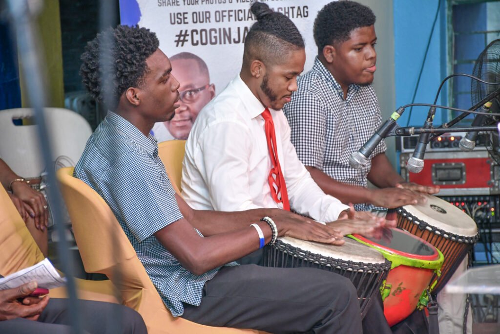 Three musicians playing hand drums at a performance, with two men wearing plaid shirts and one in a white shirt and tie.