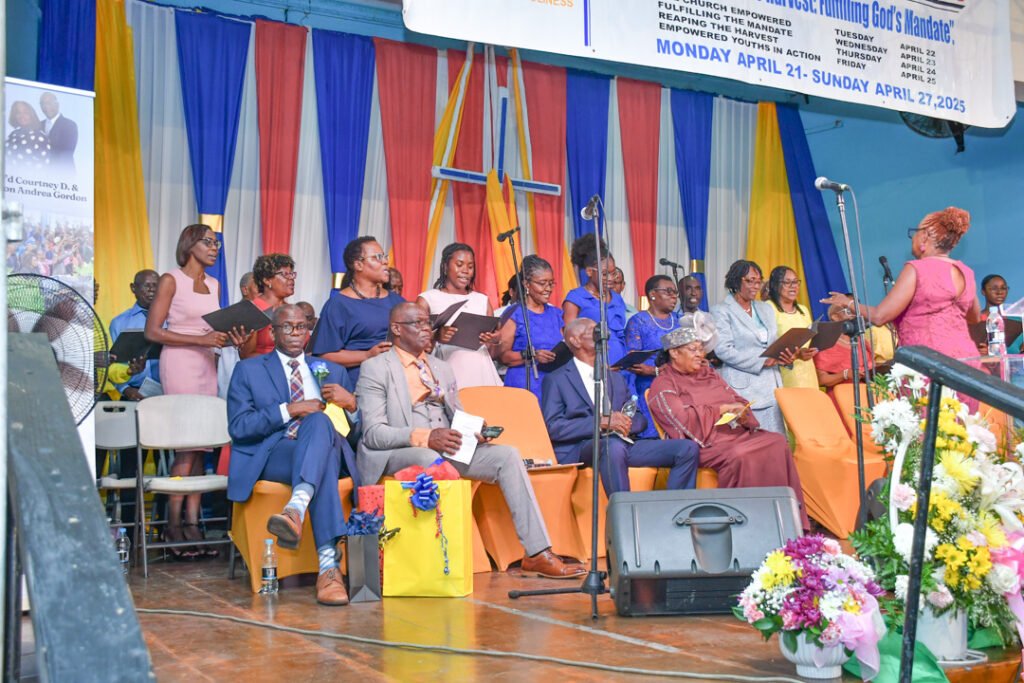A group of people, including a choir and audience members, are gathered on stage during an event, surrounded by colorful drapes and decorations. Some individuals are singing, while others are seated, listening attentively.