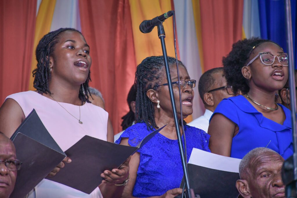 A choir performing on stage, featuring three women singing harmoniously while holding music sheets. The backdrop consists of colorful drapes.