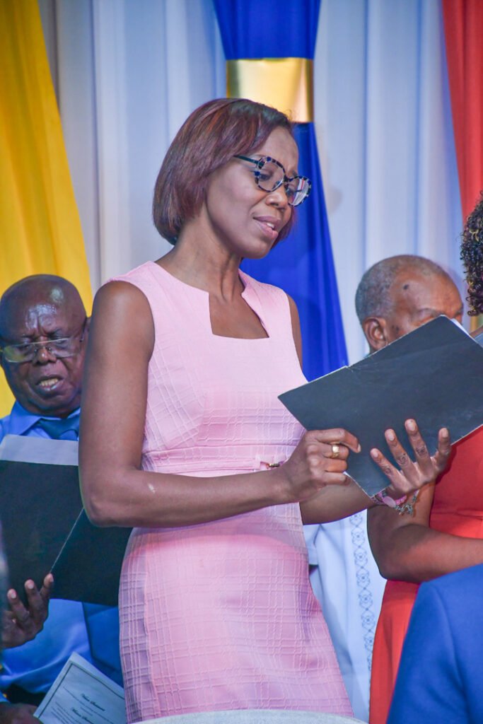 A woman in a pink dress singing or reading a sheet of music while standing in a group. In the background, other members of the group are also engaged in singing, with colorful drapes in blue and yellow behind them.