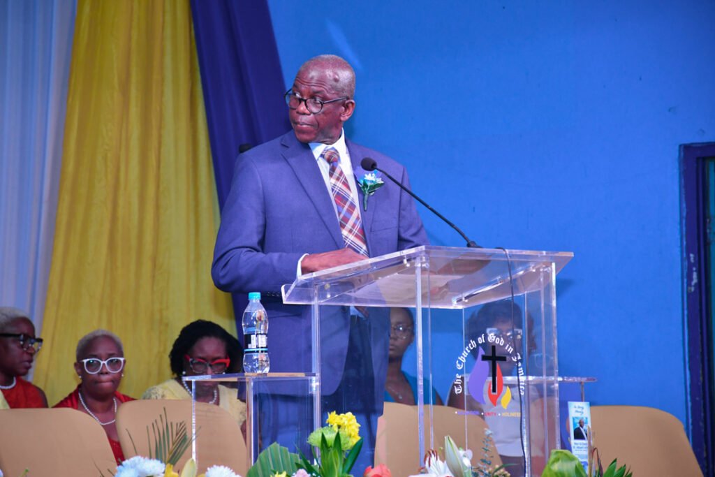 A man in a blue suit speaks at a podium during a church event, with colorful drapes in the background and an audience visible behind him.