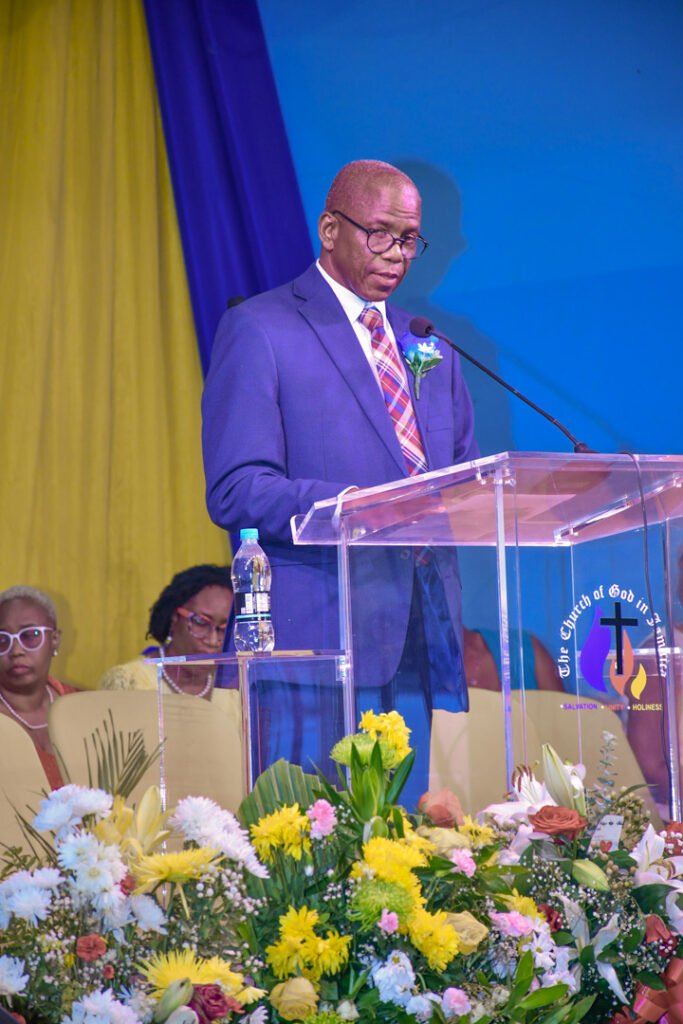 A man in a blue suit delivering a speech at a podium adorned with flowers, with attendees sitting behind him.