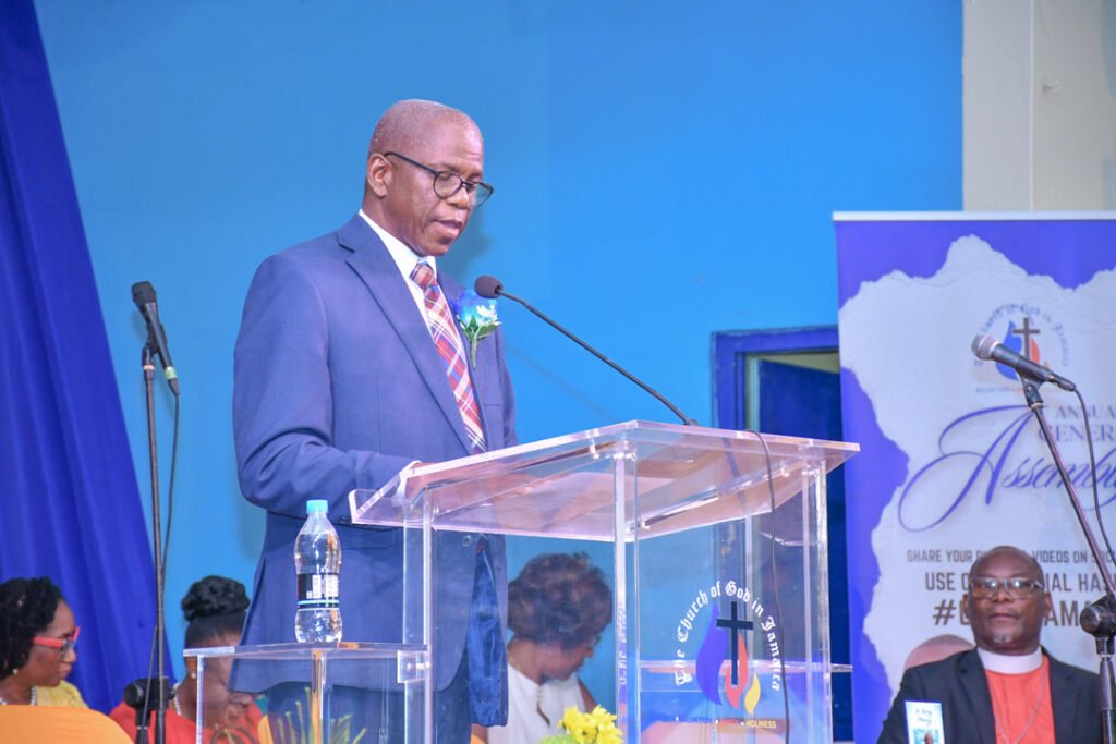 A speaker stands at a podium with a transparent lectern, addressing an audience during a formal event. The background features blue and white decor, with a banner related to a church assembly.