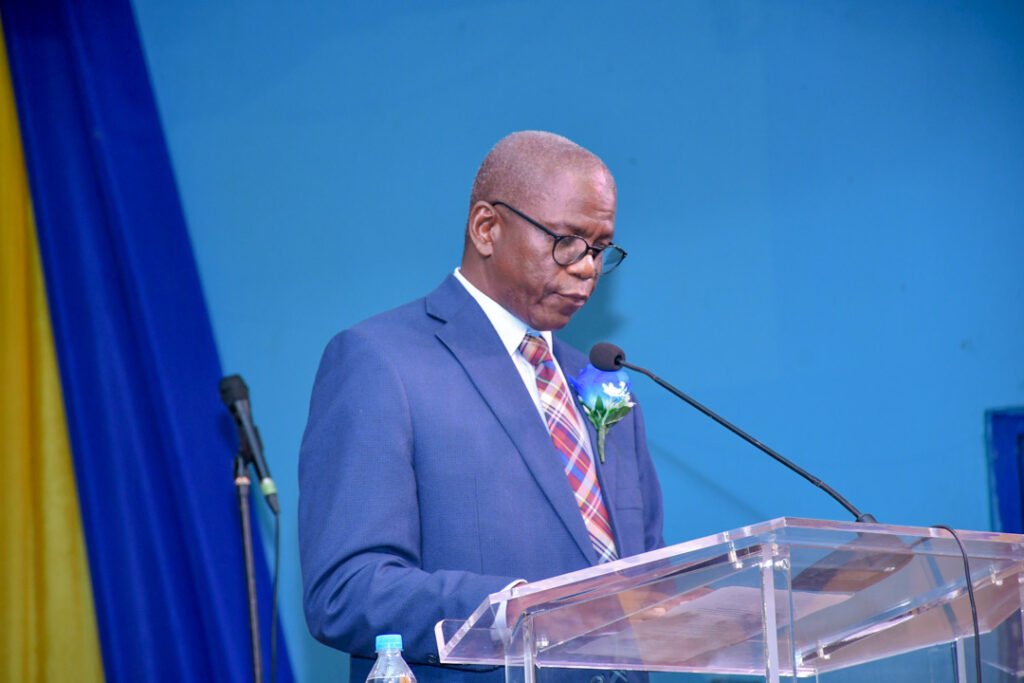 A man wearing glasses and a blue suit stands at a podium, reading from a document. He has a light blue flower pinned to his lapel, and a microphone is positioned in front of him against a blue backdrop.