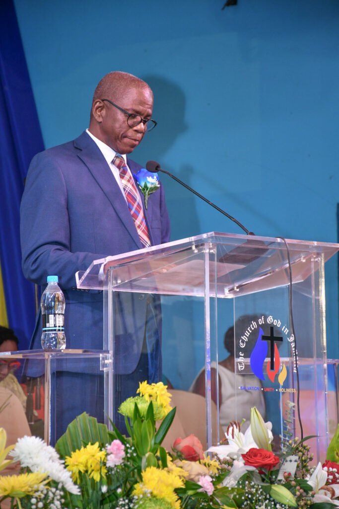 A man in a suit stands at a podium with a church logo, delivering a speech in front of colorful floral arrangements.
