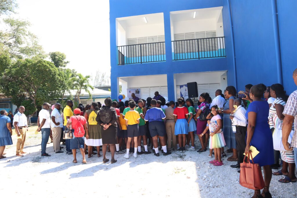 A crowd of children and adults gathered outside a blue building during a school event, with some dressed in colorful uniforms.