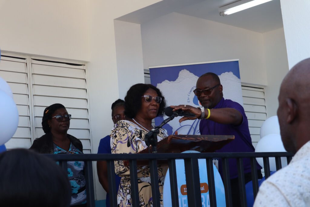 A woman speaking into a microphone at an event, with a man assisting her at the podium. Several people are visible in the background, and the setting features balloons and a banner.