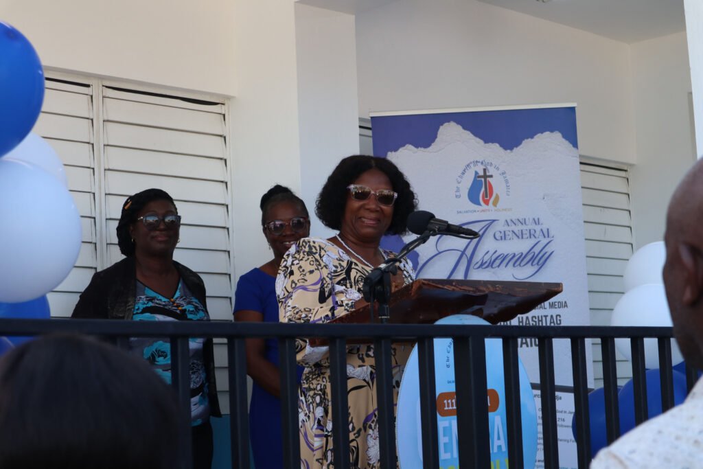 A woman speaking at a podium during an assembly, flanked by two women and decorated with blue and white balloons.