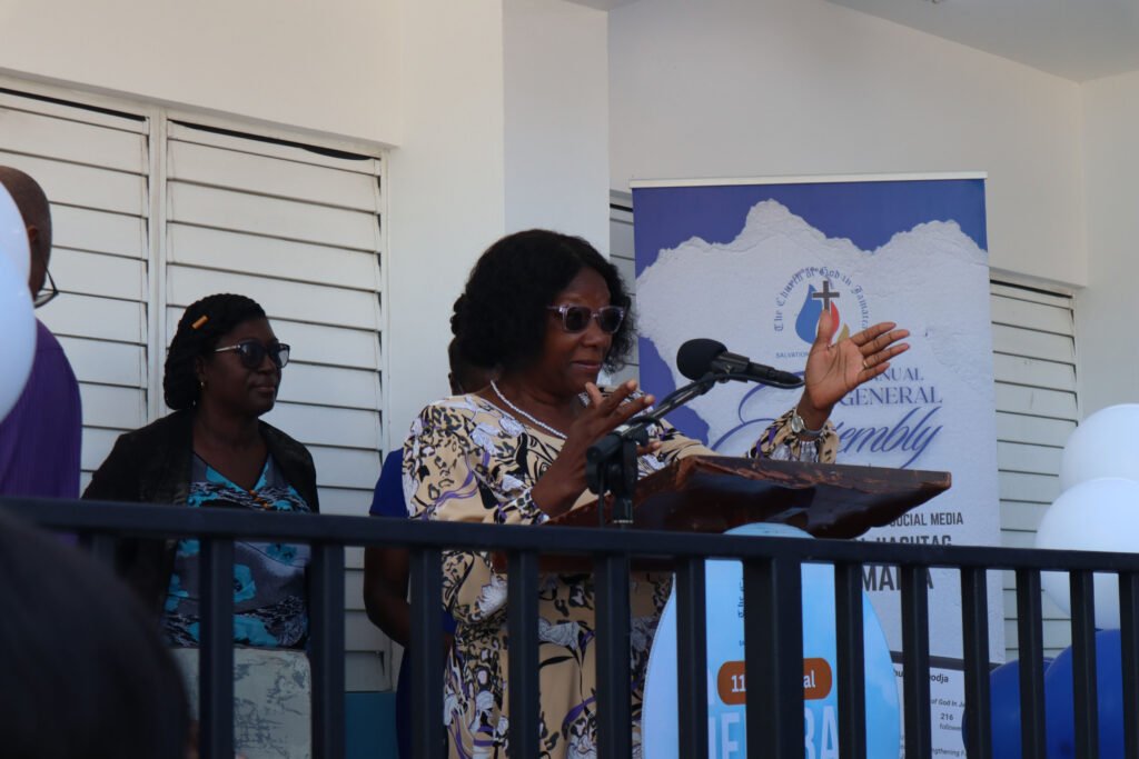 A woman speaks at a podium during a general assembly event, with a banner in the background and an audience visible.