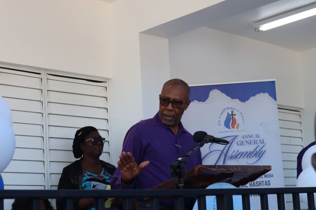 A speaker addressing an audience at an annual general assembly event, with a banner in the background displaying the event's branding.