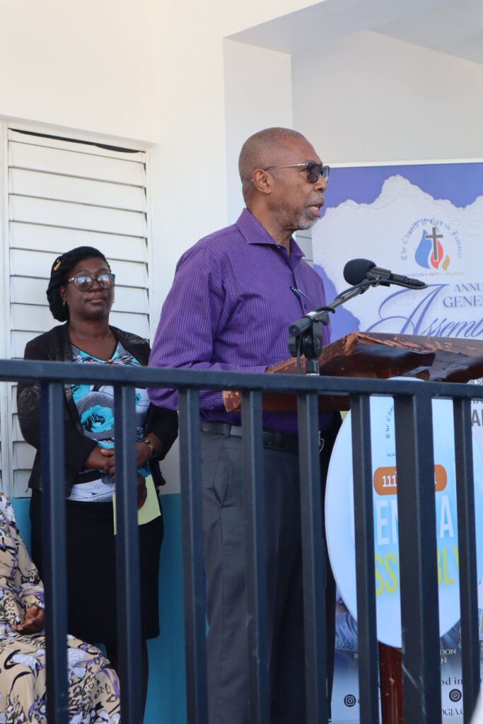 A man in a purple shirt speaking at a podium, with a woman in the background and a decorative backdrop featuring a church emblem.