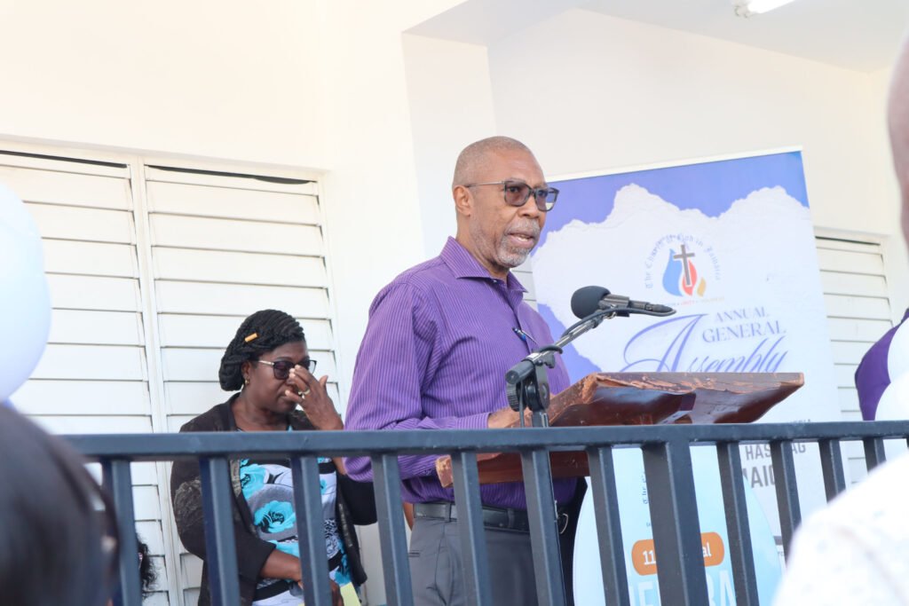 A man in a purple shirt speaks at a podium during an annual general assembly, while a woman in glasses and a black sweater is seen behind him.