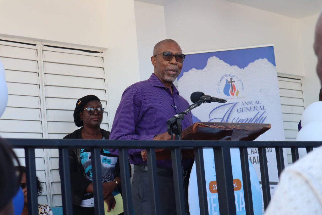 A man with glasses speaking at a podium during an event, with a woman in the background and a banner for an annual general assembly visible.