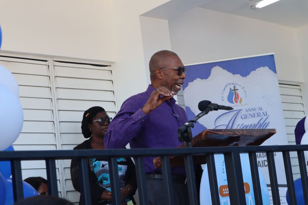 A speaker addressing an audience at an annual general assembly, with balloons and a backdrop featuring the event's branding.