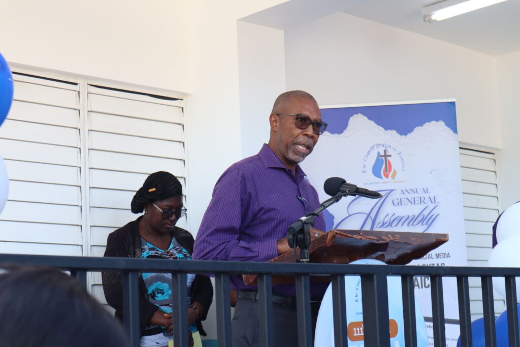 A man in glasses and a purple shirt stands at a podium speaking during an assembly event, while a woman in a black cardigan and glasses looks on.