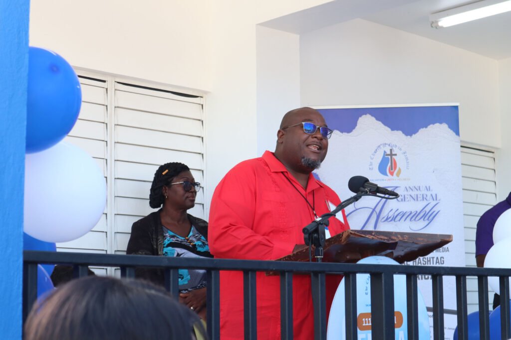 A man in a red shirt speaks at a podium during an event, while a woman in the background listens. Balloons and a banner are visible, suggesting a formal gathering.