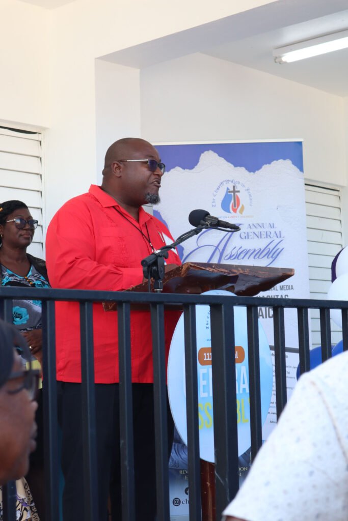 A man in a red shirt stands at a podium speaking during an annual general assembly, with attendees visible in the foreground and a banner in the background.