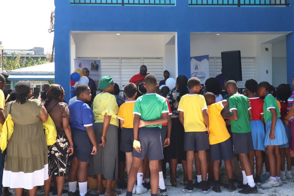 A group of children wearing colorful uniforms gather in front of a building, listening to a speaker during an event.