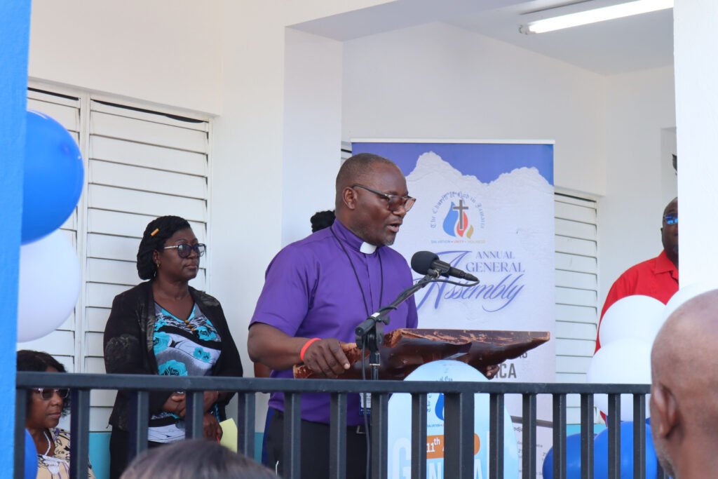 A speaker at a podium addresses an audience during an annual general assembly event. There are attendees seated with blue and white balloons in the background.