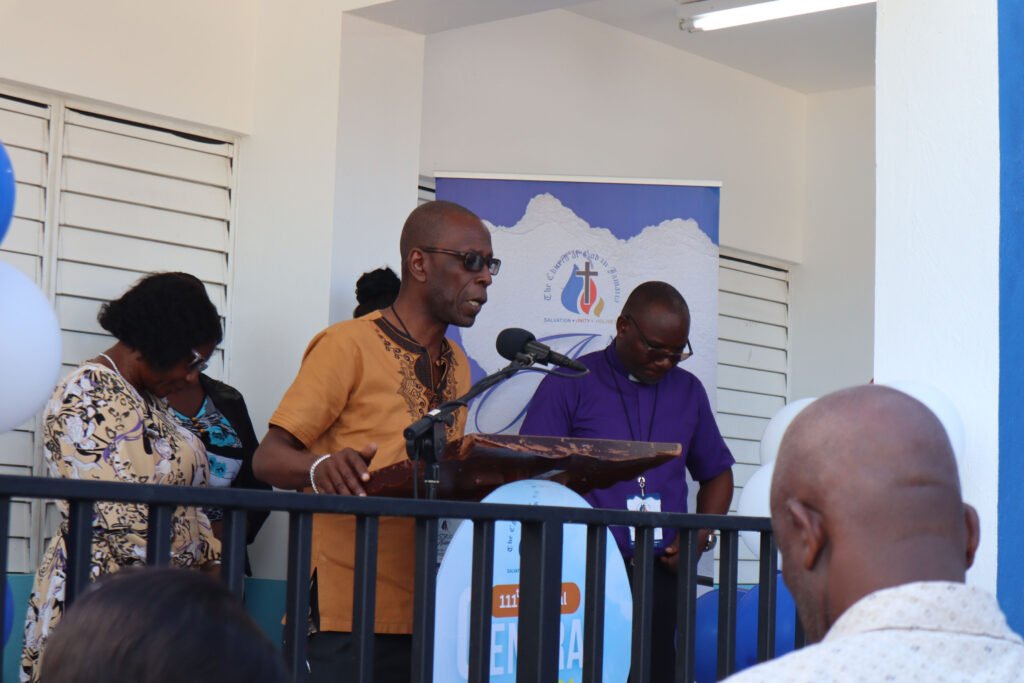 A speaker addresses an audience at a podium, with two individuals standing beside them, in front of a decorative background featuring balloons and a church logo.